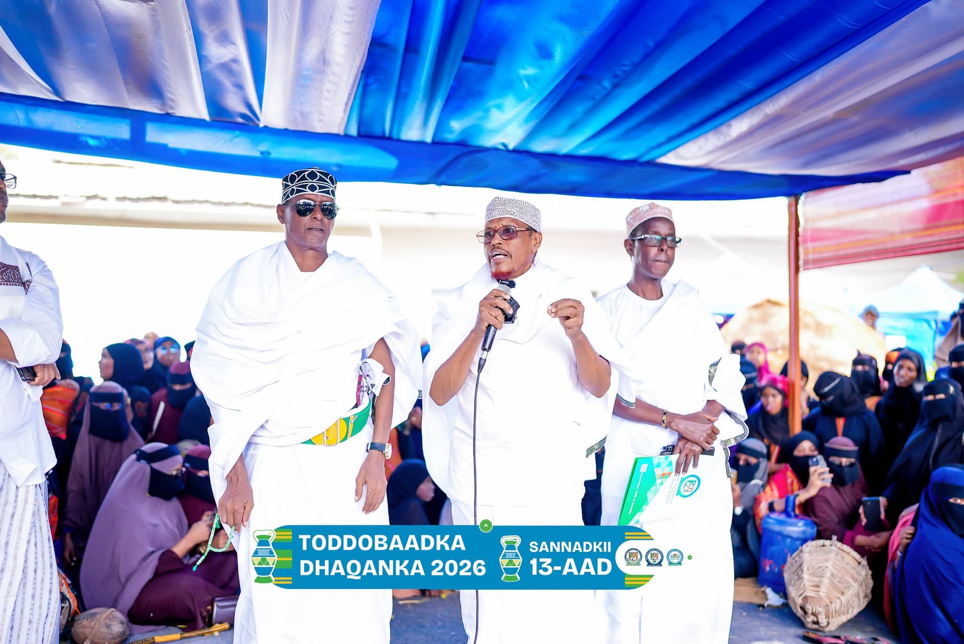 University officials in white traditional robes address a large seated crowd of students under a tent during the Cultural Week programme.
