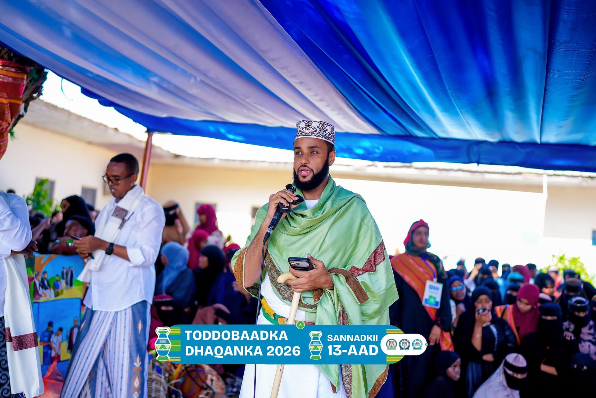 A student in traditional white and green attire speaks into a microphone before a seated audience during the Cultural Week programme.